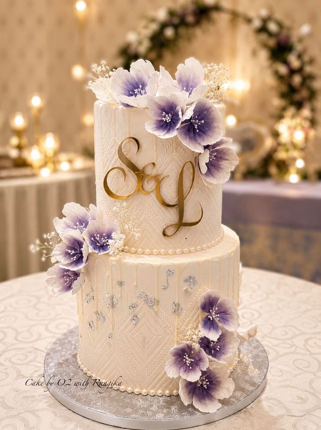 Two-tier ivory wedding cake with purple ombré sugar flowers, a gold S&L monogram and pearl trim, by Cake by O2 in Colombo