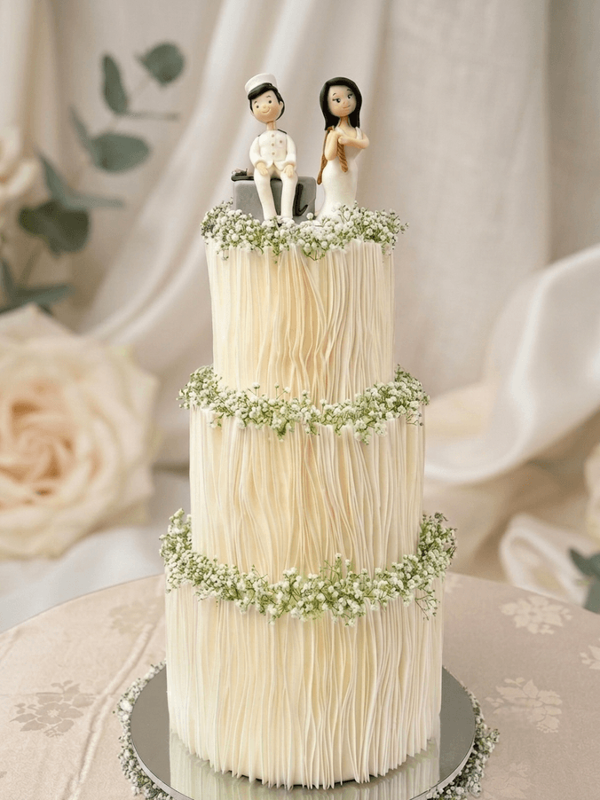 Three-tier white wedding cake with wafer-paper pleats, baby's breath garlands and a hand-modelled couple topper by Cake by O2, Colombo